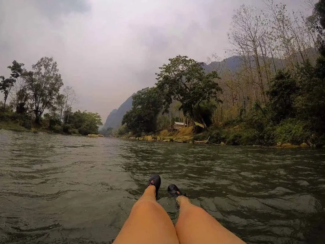 Person's perspective of their legs floating on a river with lush greenery and mountains in the background, capturing the essence of tubing in Vang Vieng