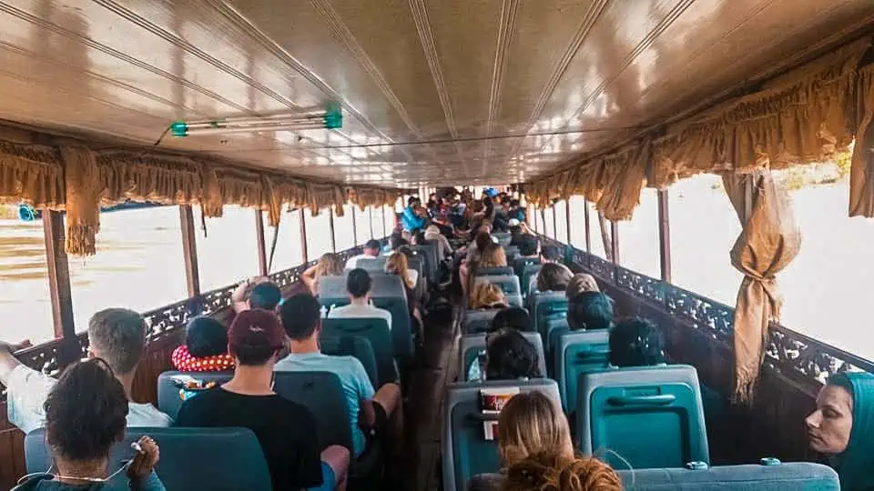 Passengers seated inside a spacious slow boat to Luang Prabang, Laos, with large windows offering a view of the Mekong River.