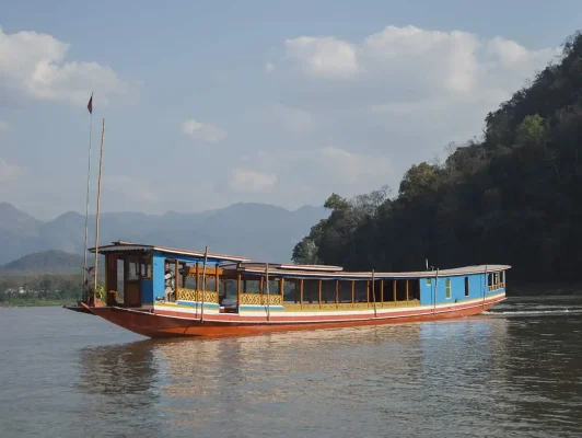 A colorful 'slow boat to Laos' cruising on the Mekong River, with a backdrop of lush green hills under a clear blue sky.