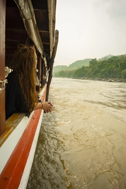 tasha amy rests her hands on the edge of a slow boat to Laos, her hair blowing in the wind as she admires the view of the Mekong River and the approaching Luang Prabang.