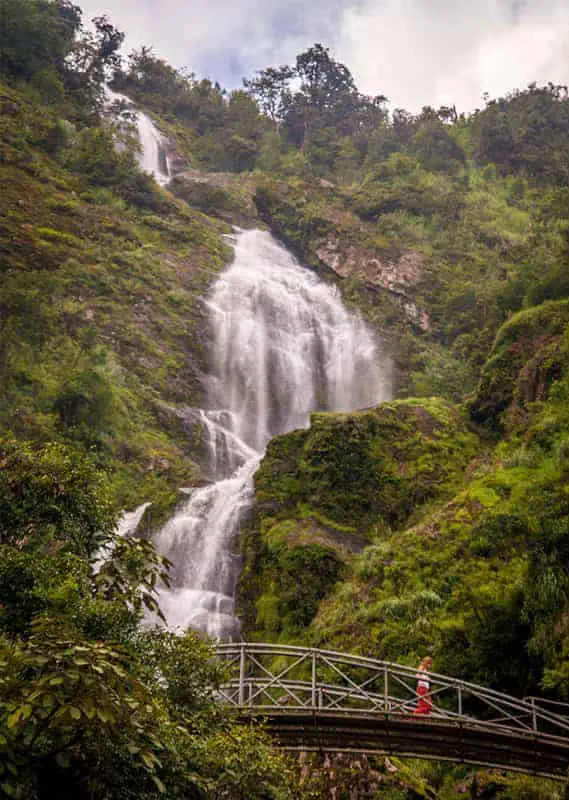 3 Days In Sapa Itinerary: Trekking, Fansipan & Homestay 49 A love waterfall drops down cliffs in Sapa, with a small bridge crossing below and a person walking across.