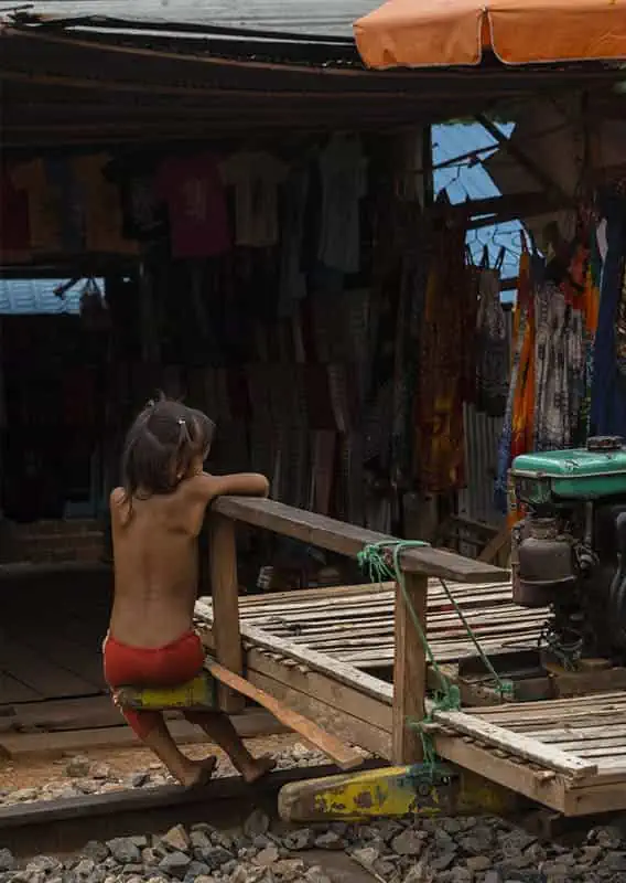 Battambang Bamboo Train: Is It Still Worth Visiting? 2026 53 A young child sits at the edge of a bamboo train cart, parked in front of a market stall with hanging clothes and in the background.