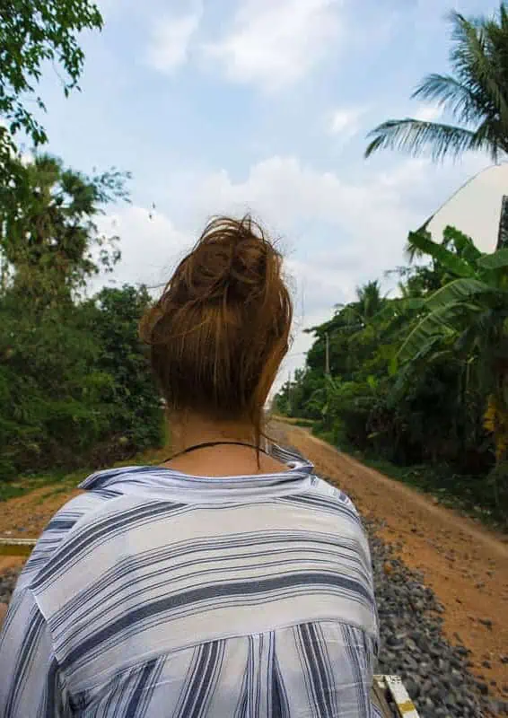Battambang Bamboo Train: Is It Still Worth Visiting? 2026 57 tasha amy with her back to the camera sits on a battambang bamboo train riding along the railway, surrounded by palm trees and jungle.