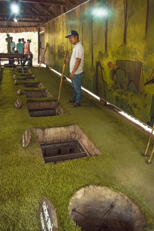 Best Cu Chi Tunnels Tours: Cheap VS Expensive 2026 52 An interior view of a Cu Chi Tunnels exhibit with various open trapdoors in the ground, demonstrating guerrilla tactics with a guide and visitors in the background, painted walls depict a jungle environment.