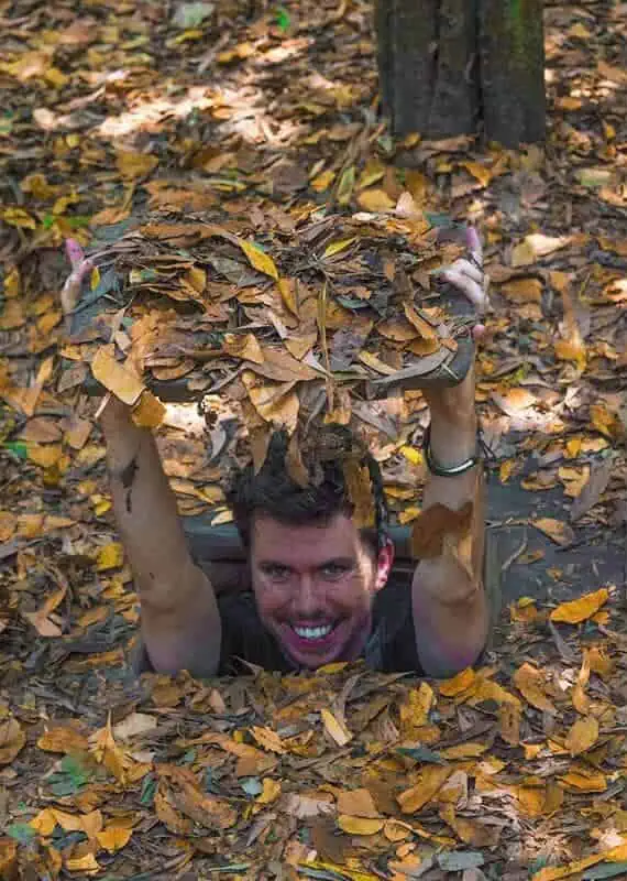Best Cu Chi Tunnels Tours: Cheap VS Expensive 2026 48 A man smiling joyfully while emerging from a Cu Chi Tunnel camouflaged with a cover of dry leaves, with the dense forest around him.
