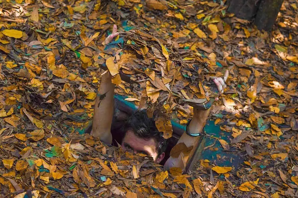 Best Cu Chi Tunnels Tours: Cheap VS Expensive 2026 51 A man peers out of a Cu Chi tunnel, concealed by a camouflage of dry leaves, illustrating the stealthy design of the tunnels.