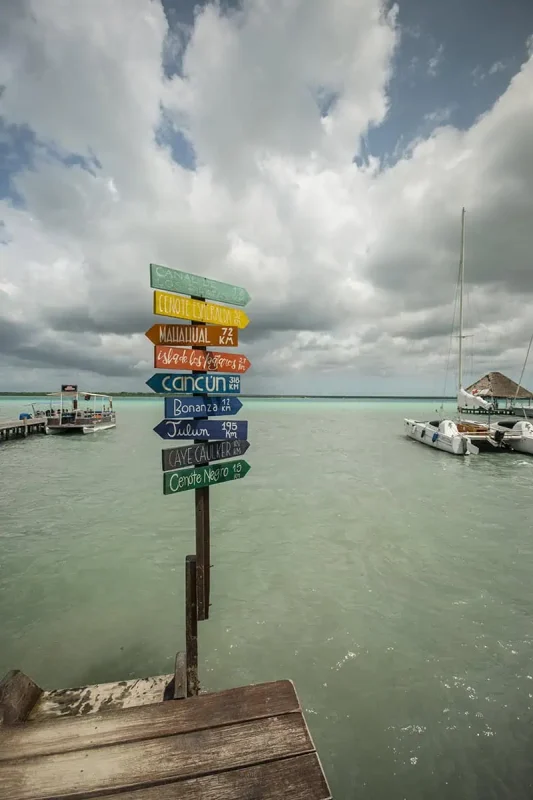 Mexico Belize Guatemala Itinerary: BEST Backpacking Route 70 A colorful signpost on a wooden dock in Bacalar pointing to various destinations including Cancun, Cenote Esmeralda, and Caye Caulker. Each arrow is painted in a different vibrant color, adding a playful touch to the dock scene with a backdrop of cloudy skies and calm turquoise waters.