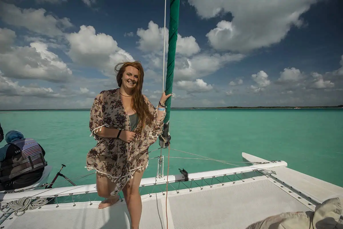 Mexico Belize Guatemala Itinerary: BEST Backpacking Route 72 tasha amy board a sailboat on the bright turquoise waters of Bacalar. She is leaning on the netting of the catamaran, dressed in a floral dress, with her hair blowing in the wind. The sky above is partly cloudy, emphasizing the vivid color of the water.