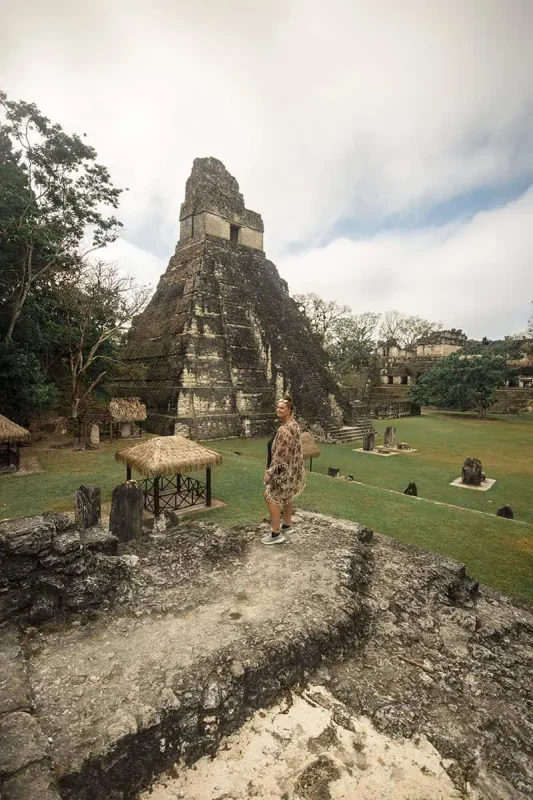 Mexico Belize Guatemala Itinerary: BEST Backpacking Route 94 tasha amy standing in front of a massive Mayan temple at Tikal, part of a historical site near Flores. The temple rises dramatically against a backdrop of lush greenery and overcast skies
