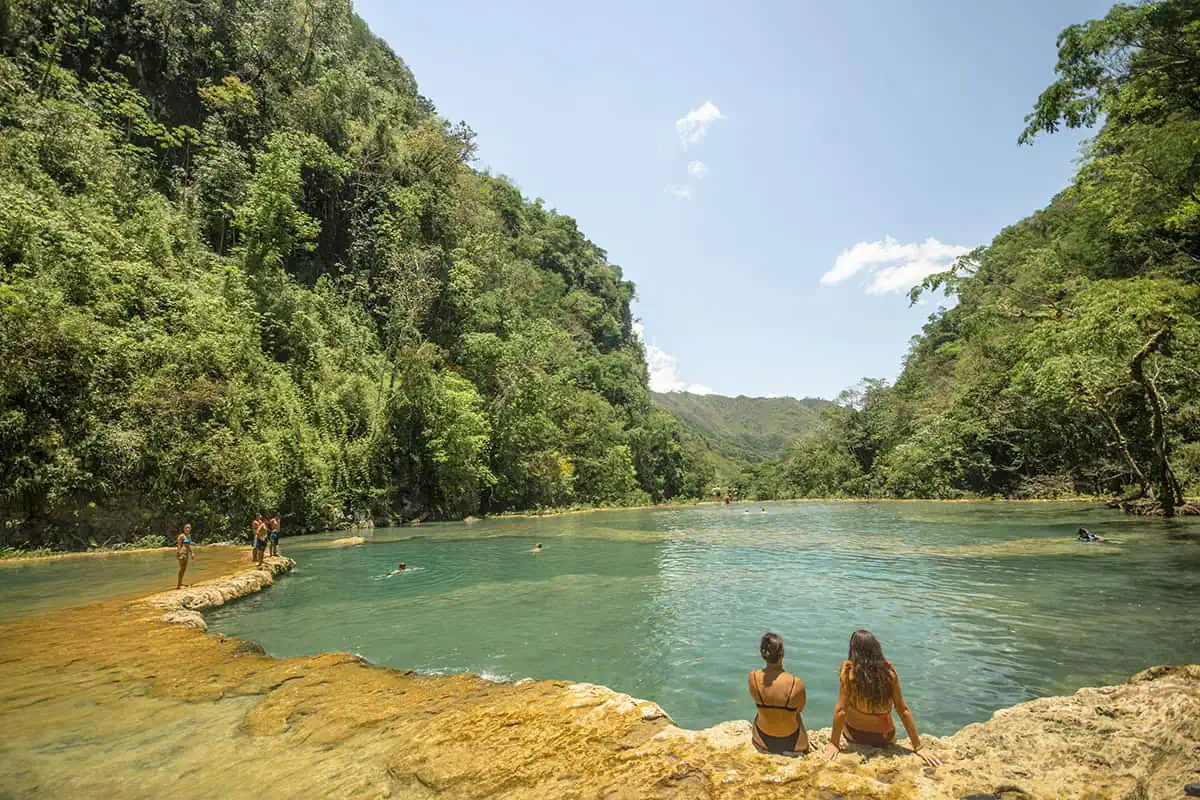 Mexico Belize Guatemala Itinerary: BEST Backpacking Route 104 An idyllic view of Semuc Champey's natural limestone pools, nestled in a lush jungle setting in Guatemala. The vibrant turquoise waters are surrounded by rich green vegetation and a rugged mountain backdrop. People enjoy the serene environment, swimming and relaxing along the water's edge