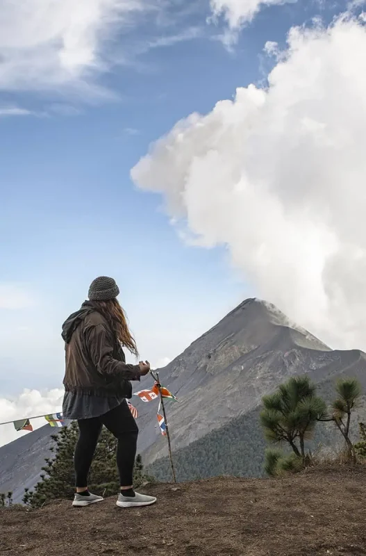 Mexico Belize Guatemala Itinerary: BEST Backpacking Route 110 tasha amy standing on a mountain peak, observing a nearby active volcano emitting a plume of smoke in Antigua. She is dressed in outdoor gear and holding colorful prayer flags, which add a vivid touch against the rugged volcanic backdrop. This adventurous experience is a notable highlight in the Mexico, Belize, Guatemala itinerary