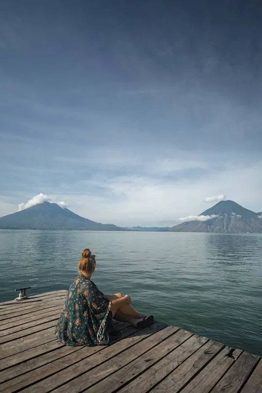 Mexico Belize Guatemala Itinerary: BEST Backpacking Route 116 tasha amy sits on a wooden dock overlooking Lake Atitlán, surrounded by majestic volcanoes under a clear blue sky. Her contemplative pose captures a moment of peace as she gazes out over the calm waters, framed by the natural beauty of Guatemala. This picturesque setting is a serene highlight on the Mexico, Belize, Guatemala itinerary.
