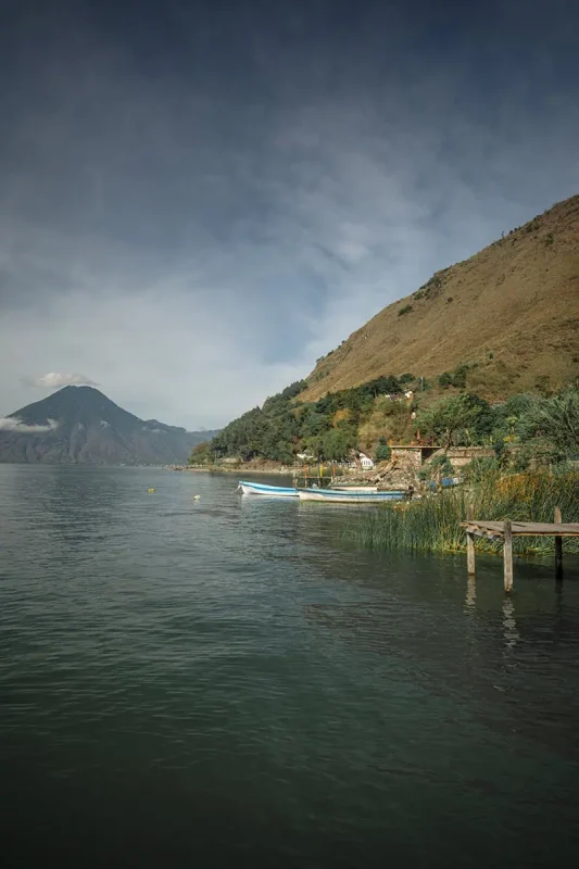 Mexico Belize Guatemala Itinerary: BEST Backpacking Route 117 A tranquil scene at Lake Atitlán with several small boats moored along a wooden pier, set against a backdrop of lush green hills and a calm lake surface. The clear skies and peaceful water reflect the serene and untouched nature of the area, inviting exploration and relaxation.