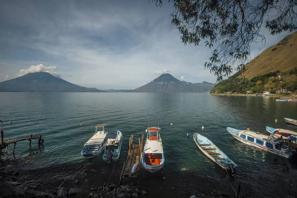 Mexico Belize Guatemala Itinerary: BEST Backpacking Route 118 A panoramic view of Lake Atitlán, captured from a lakeside spot where boats are tied up to a rustic wooden dock. The lake is flanked by towering volcanoes and hills, with trees overhanging the water's edge, presenting a perfect blend of natural elements and serene landscapes.