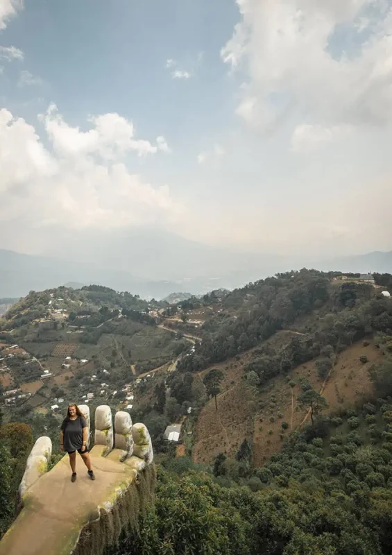 Mexico Belize Guatemala Itinerary: BEST Backpacking Route 109 tasha amy stands on a uniquely designed viewpoint that resembles a hand, located on the edge of a hill in Antigua. Below, a panoramic view of a lush valley dotted with small buildings and patches of agriculture extends into the distance, framed by a sky filled with light clouds.
