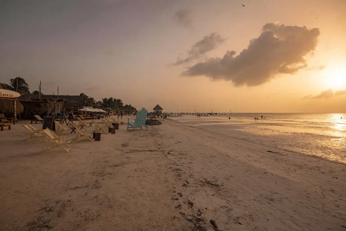 Mexico Belize Guatemala Itinerary: BEST Backpacking Route 56 A tranquil sunset view at Isla Holbox, featuring a softly lit sandy beach with scattered wooden chairs and tables. Several beachside bars and a wooden pier stretch into the calm sea, under a sky painted with hues of orange and pink. A single cloud floats above the horizon in an otherwise clear sky