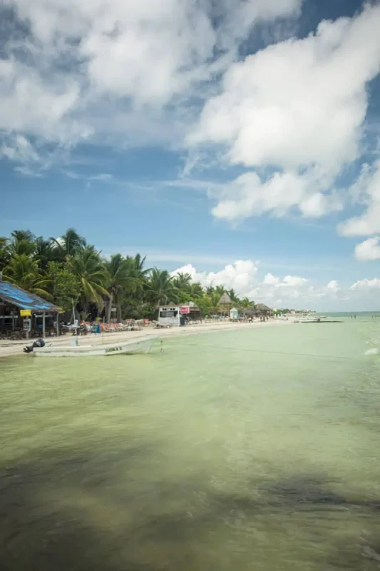 Mexico Belize Guatemala Itinerary: BEST Backpacking Route 55 Crystal-clear turquoise waters edge a vibrant tropical beach at Isla Holbox. The shore is lined with palm trees and casual beachfront eateries under a bright blue sky with fluffy clouds. A few boats are moored in the shallow water, adding to the serene tropical atmosphere.