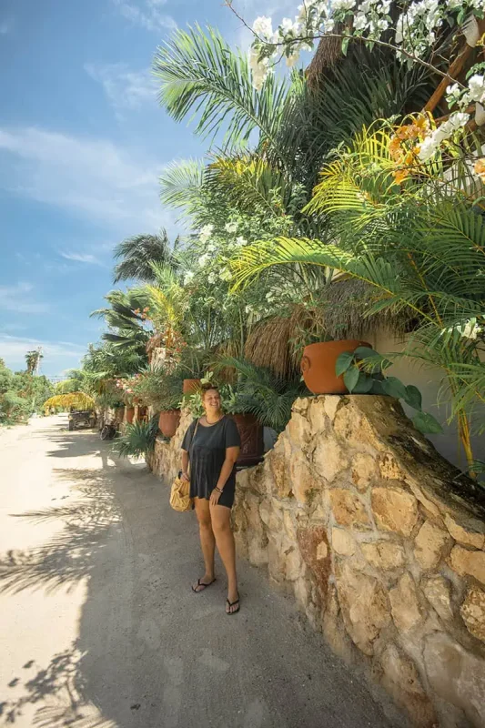 Mexico Belize Guatemala Itinerary: BEST Backpacking Route 54 tasha amy in casual summer attire stands on a sandy pathway surrounded by lush tropical greenery and flowering plants on Isla Holbox. The path is flanked by stone walls and vibrant, densely packed tropical plants, providing a rich, natural environment.