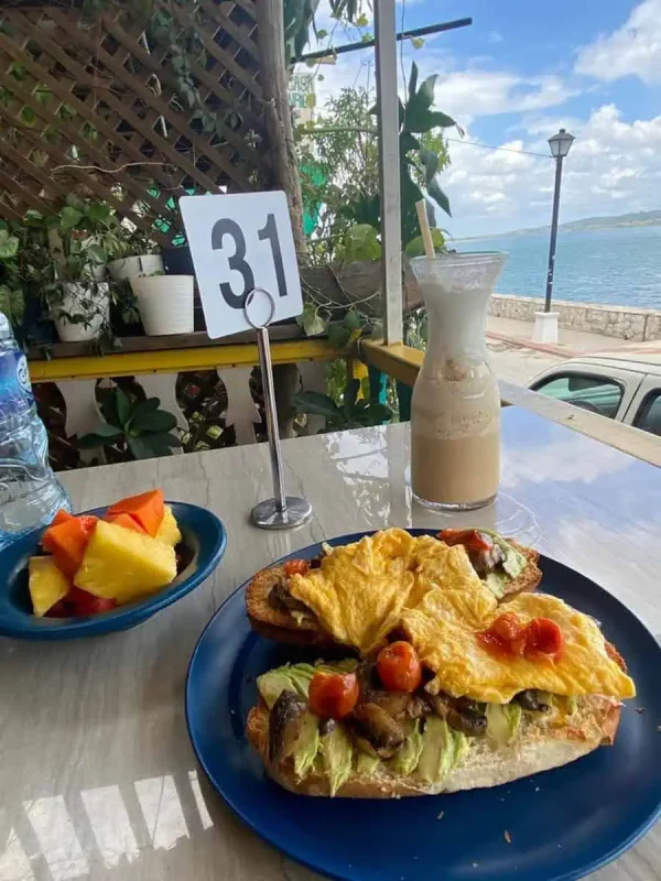 Mexico Belize Guatemala Itinerary: BEST Backpacking Route 93 A meal set up on a beachfront restaurant table in Flores, including a hearty open sandwich topped with avocado, scrambled eggs, and tomatoes on crusty bread. A glass of iced coffee and a bowl of fresh fruit salad complement the meal, set against the backdrop of a serene ocean view, making it part of a memorable Mexico, Belize, Guatemala itinerary.