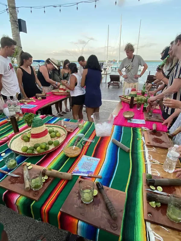 Mexico Belize Guatemala Itinerary: BEST Backpacking Route 77 vibrant mojito-making class by the sea in Bacalar, Mexico. A group of tourists, of various ages and backgrounds, is gathered around a colorful table covered with a traditional Mexican "sarape" tablecloth. They're actively engaged in learning how to make mojitos, using ingredients like fresh limes and mint, displayed on rustic wooden chopping boards. The scene is lively and festive, set against a backdrop of sailboats moored in the turquoise waters of Bacalar Lagoon, enhancing the tropical and social atmosphere of the experience.