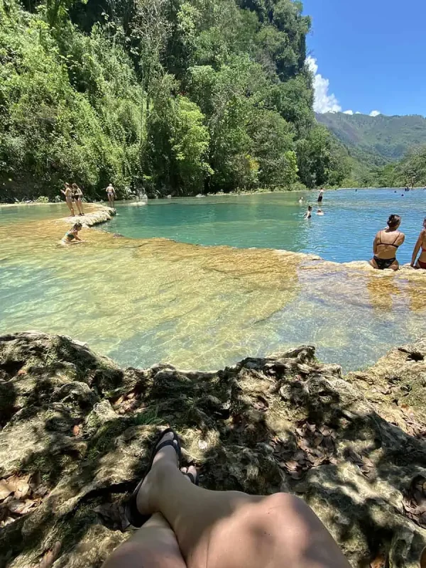 Mexico Belize Guatemala Itinerary: BEST Backpacking Route 102 A first-person perspective showing a woman's legs as she sits on the rocky edge of a natural pool in Semuc Champey. The clear green waters stretch out in front of her, bordered by lush greenery and a vibrant forest scene, reflecting a peaceful and relaxing atmosphere.