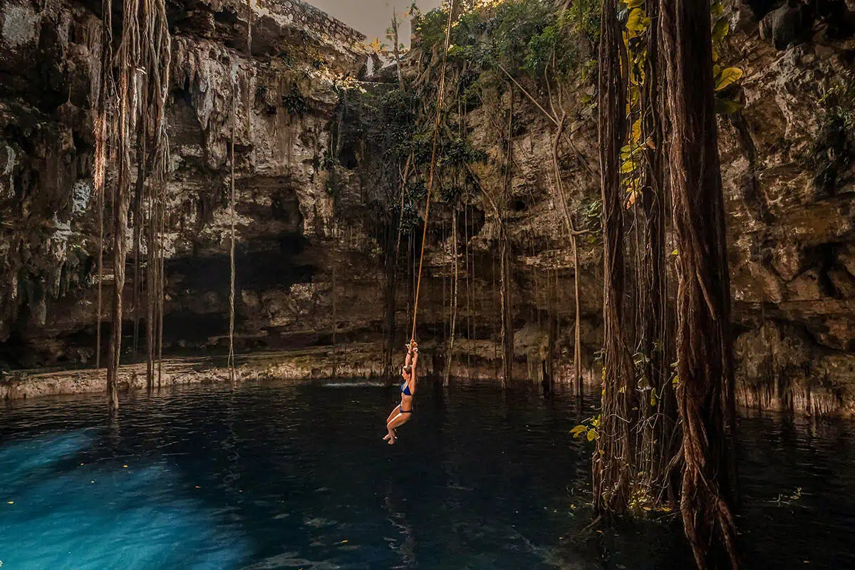 Mexico Belize Guatemala Itinerary: BEST Backpacking Route 5 A breathtaking scene of a woman swinging on a rope over a cenote in Tulum, Mexico. This natural sinkhole is surrounded by rugged cliffs adorned with hanging roots and lush vegetation. The water reflects a deep, vivid blue hue, creating a serene yet adventurous atmosphere. this is a must on the mexico belize guatemala itinerary.