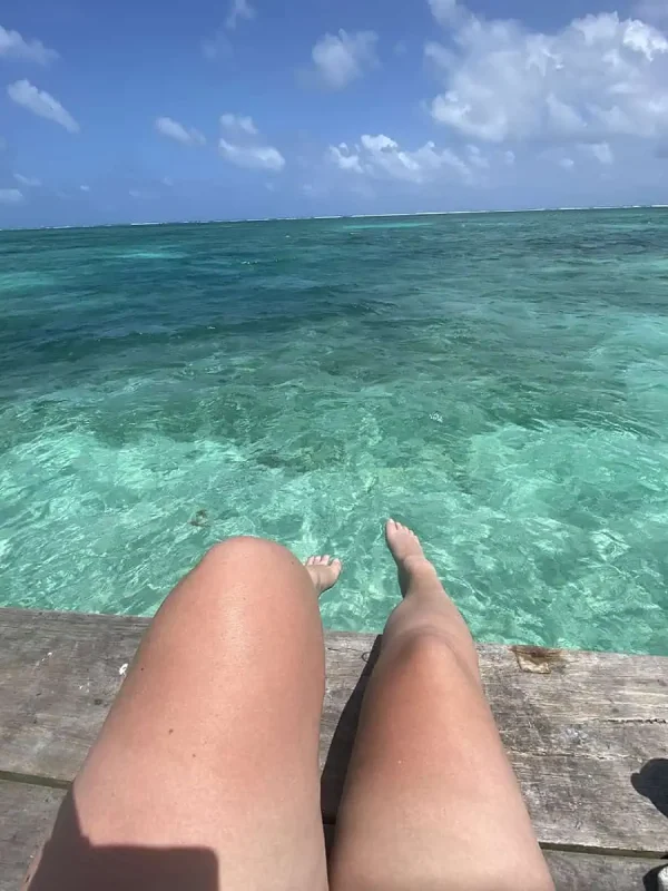Bacalar To Caye Caulker: The ULTIMATE Travel Guide 2026 65 View of a Tasha's legs dangling off a wooden dock, with her feet hovering above the clear waters of Caye Caulker.