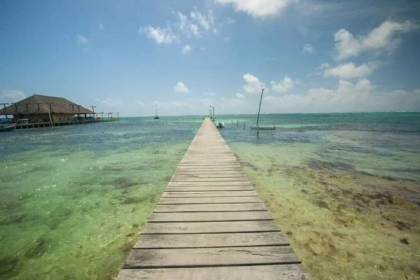 A wooden pier extends into the calm, clear turquoise waters of Belize, leading to a thatched-roof structure at the end. The sunny sky and a few moored sailboats create a serene setting showcasing belize in april