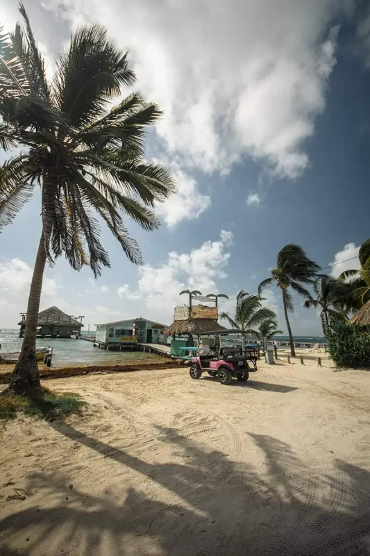 Belize In April: What You NEED To Know 57 A quaint sandy path runs along a serene beach in Belize, flanked by rustic buildings and thatched-roof bars. A pink golf cart parked on the side adds a touch of modern convenience to this otherwise natural and tranquil setting.