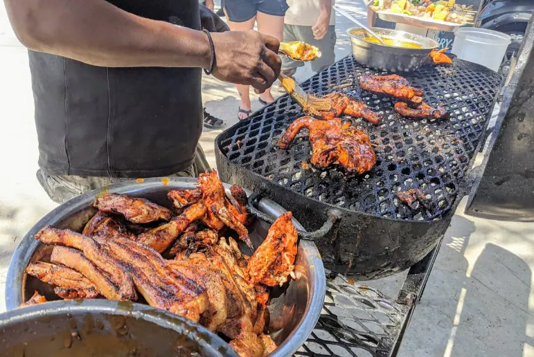 BEST Restaurants On Caye Caulker Worth Trying 2026 2 A bustling street food scene at Chef Kareem's UnBelizeable Lunch, known as one of the best restaurants on Caye Caulker for authentic local cuisine. The photo shows a chef, deftly handling various pieces of juicy, barbecued chicken and ribs over a grill, with a bowl of marinade at hand for basting. The grill is filled with deliciously charred meats, enticing passersby with its smoky aroma. This image captures the vibrant street food culture that is a hallmark of Caye Caulker’s culinary offerings.