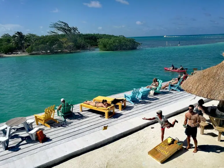 A vibrant, sunny day at Lazy Lizard, one of the best restaurants on Caye Caulker, captured from a bustling wooden pier. The image showcases a group of people enjoying the clear turquoise waters and serene views. Several lounge chairs and colorful wooden benches provide ample seating for guests to relax and soak in the sun. A few individuals are engaging in casual activities, adding to the lively atmosphere of this popular waterside destination.