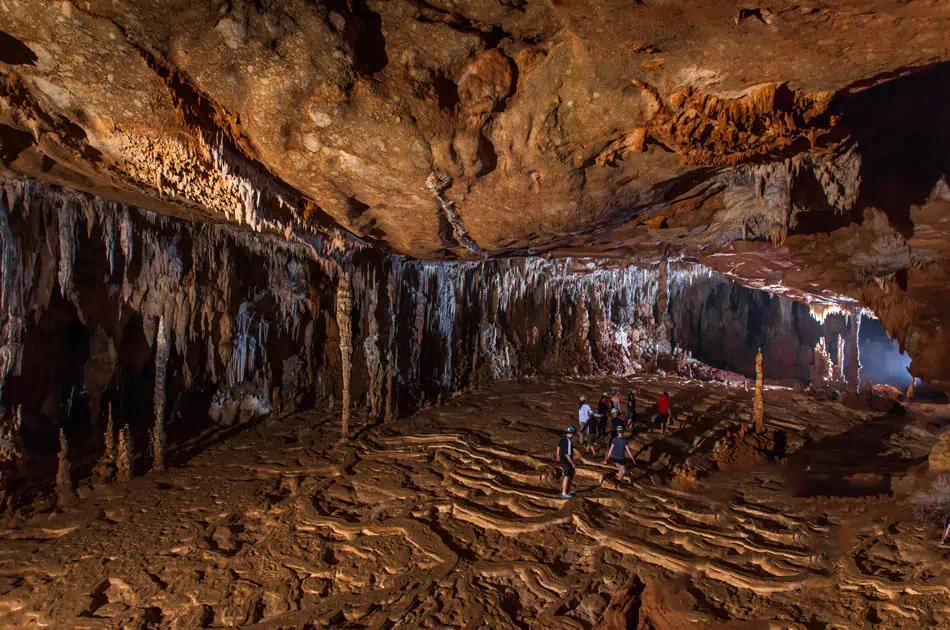 Best Things To Do In San Ignacio: Caves, Ruins & Day Trips 76 Interior of the ATM cave featuring intricate stalactites hanging from the ceiling and stalagmites rising from the floor. A group of tourists, equipped with helmets and lights, explore the vast underground formations, showcasing one of the popular adventure activities in San Ignacio.