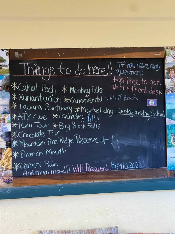 Best Things To Do In San Ignacio: Caves, Ruins & Day Trips 82 Chalkboard in a hostel listing various activities under 'Things to do here!!' including Cahal Pech, Xunantunich, Iguana Sanctuary, ATM Cave, and more, highlighting numerous attractions and activities in San Ignacio.