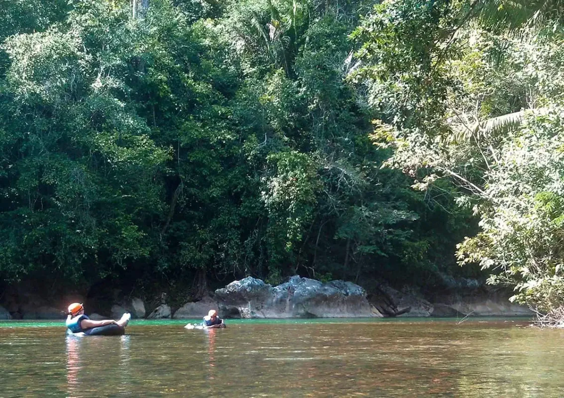 Best Things To Do In San Ignacio: Caves, Ruins & Day Trips 61 Two people floating on inner tubes in a calm river surrounded by dense jungle in San Ignacio. The participants are wearing life vests and helmets, enjoying the peaceful tubing experience as one of the adventurous things to do in San Ignacio.