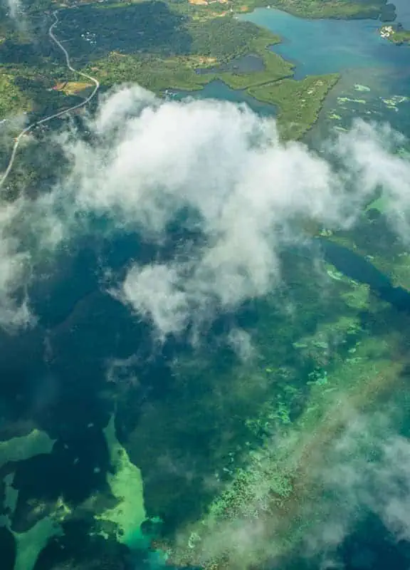 Cancun To Caye Caulker: The 4 Easiest Ways To Travel 62 A stunning aerial view from a plane window revealing the vibrant turquoise waters of the Caribbean Sea surrounding the coastline of Caye Caulker, Belize. The foreground shows the plane's metallic railing, while below, dense mangrove forests, a winding road, and a sandy shoreline characterize the island. This captivating sight greets travelers flying from Cancun to Caye Caulker, offering a glimpse of the paradise below