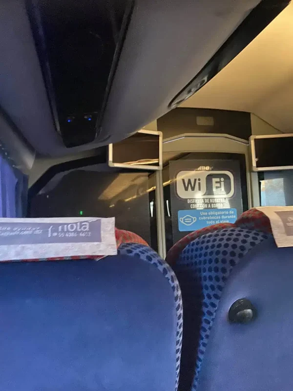 Cancun To Caye Caulker: The 4 Easiest Ways To Travel 58 Interior of an ADO bus traveling from Cancun to Caye Caulker. The bus features seat-back displays and Wi-Fi signs visible on the rear door, offering connectivity during the journey. The shot is taken from a passenger's perspective, showing blue headrests and rows of seats.
