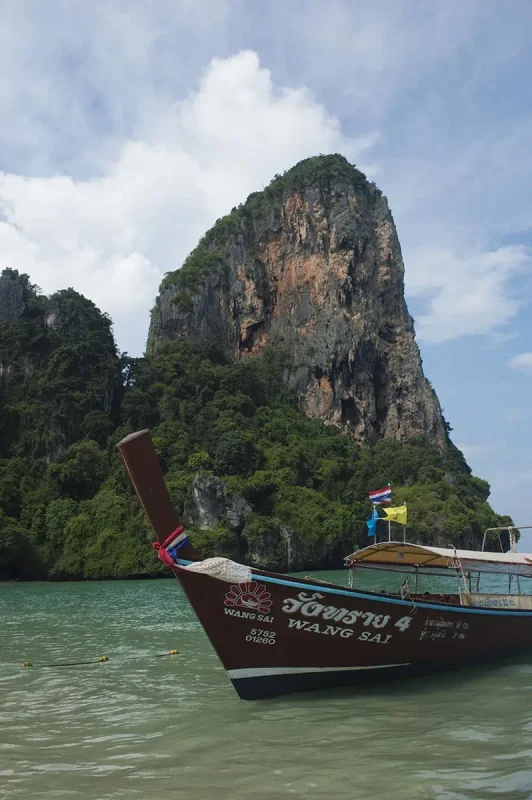Is Railay Beach Closed: What You Need To Know (2026) 50 A long-tail boat named "Wang Sai" anchored near a towering limestone cliff covered in greenery at Railay Beach. The presence of boats and tourists hints at the beach being operational