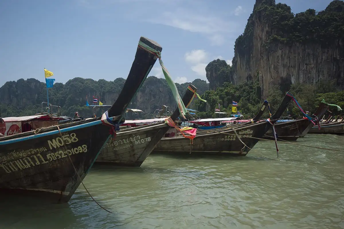 Is Railay Beach Closed: What You Need To Know (2026) 57 Several long-tail boats with colorful flags are lined up in the water with towering cliffs in the background at Railay Beach. This busy scene implies active tourist engagement, questioning if Railay Beach is closed.