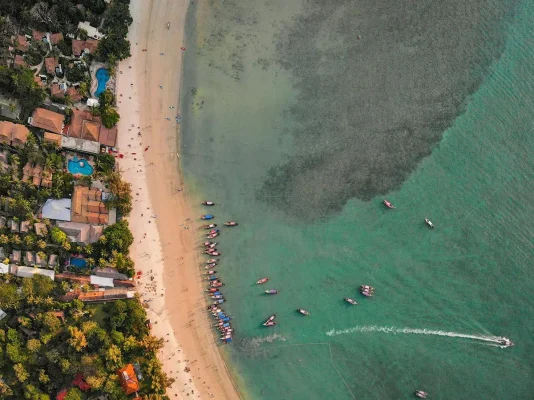 Aerial view of Railay Beach showcasing its sandy shore lined with boats, turquoise waters, and surrounding lush greenery. Though with the latest island shutdown is railay beach closed