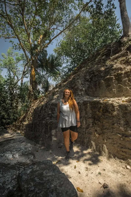 Mexico And Belize Itinerary: From 2 to 5 Weeks 99 tasha amystanding and smiling on a shaded path beside ancient Mayan ruins surrounded by dense tropical forest. She is wearing a sleeveless top and shorts, capturing the essence of adventure and exploration in belize.