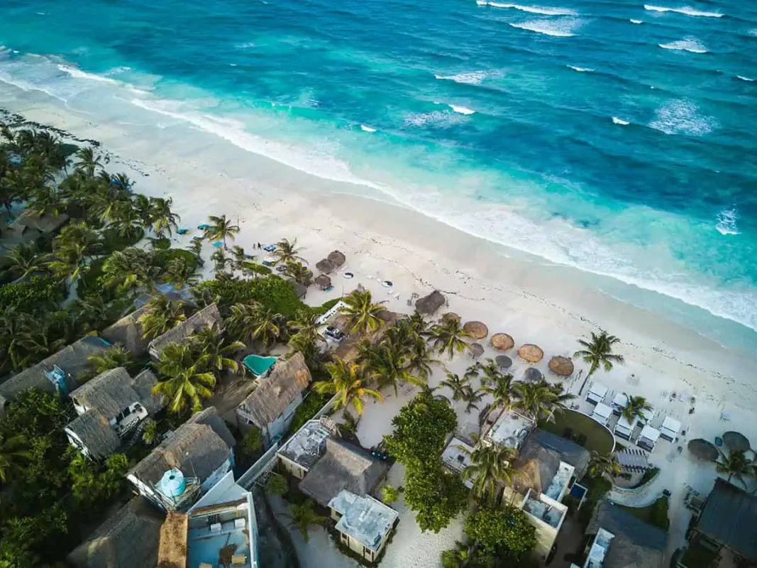 Mexico And Belize Itinerary: From 2 to 5 Weeks 1 Aerial view of a picturesque beach in Mexico with clear turquoise waters and gentle waves meeting a pristine sandy shore, the perfect place to visit on a mexico and belize itinerary. The beach is lined with palm trees and several thatched-roof huts, some with small swimming pools, offering a tranquil and inviting tropical getaway.
