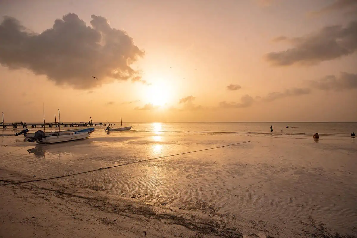 Mexico And Belize Itinerary: From 2 to 5 Weeks 57 Sunset on an isla holbox beach with small boats anchored in shallow water. The sky is painted with warm hues of orange and pink, reflecting on the water, creating a serene and picturesque moment.