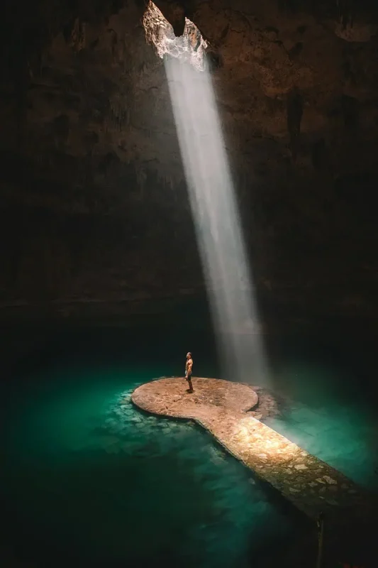 Mexico And Belize Itinerary: From 2 to 5 Weeks 73 A person standing on a stone platform inside a dark cenote, illuminated by a dramatic beam of sunlight from an opening above. The clear turquoise water surrounding the platform adds to the mystical and awe-inspiring atmosphere of this natural wonder.