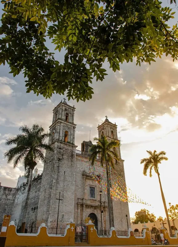 Mexico And Belize Itinerary: From 2 to 5 Weeks 64 Historic cathedral with twin bell towers framed by palm trees and lush green leaves in Valladolid, Mexico. The late afternoon sun casts a warm glow on the stone facade, capturing the architectural beauty and cultural richness of the region.