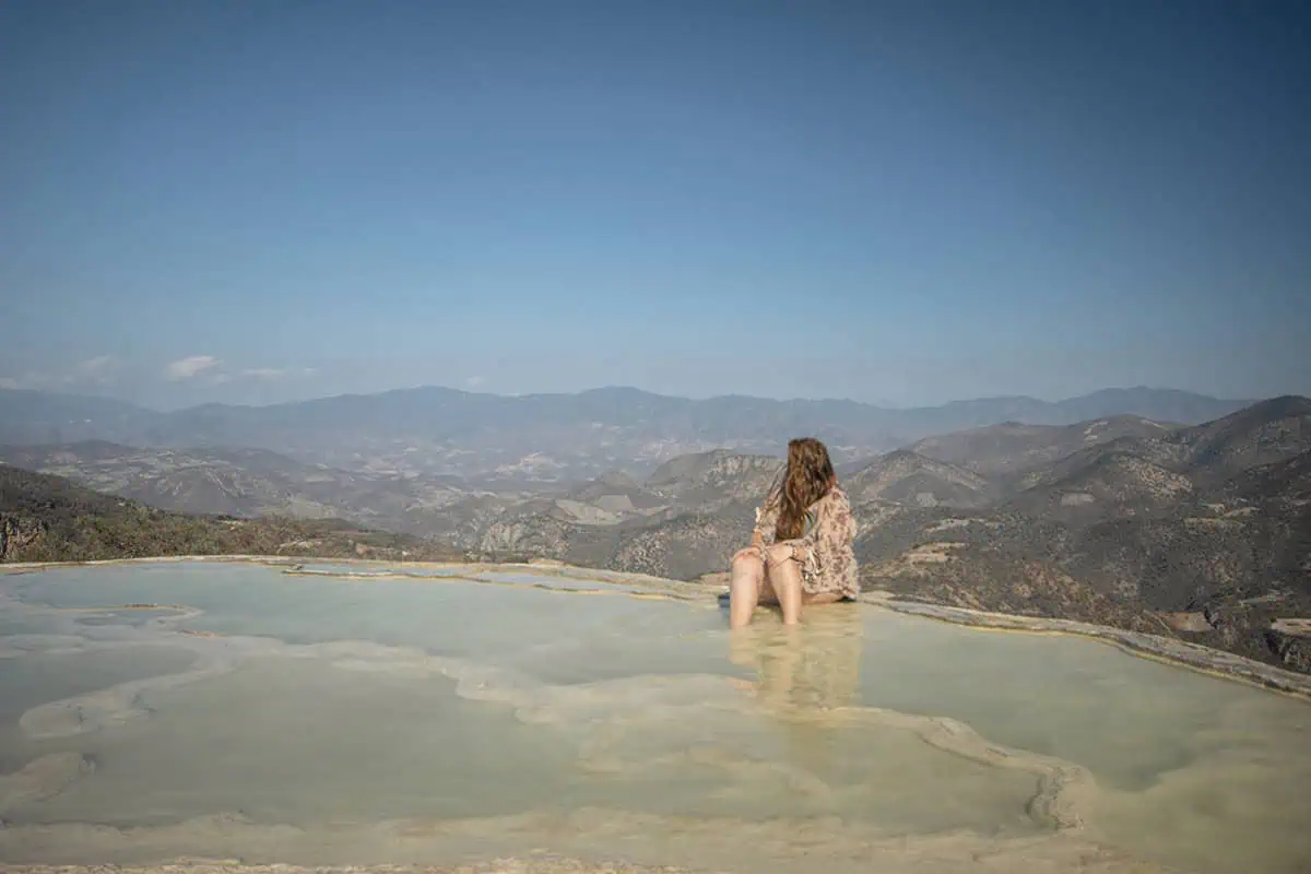 Mexico And Belize Itinerary: From 2 to 5 Weeks 101 Tasha Amy sitting at the edge of a natural infinity pool, dipping her feet into the water while gazing at the expansive mountainous landscape. The clear sky and distant hills provide a breathtaking view.