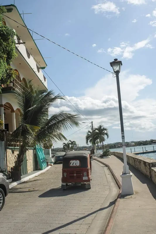 Travel San Ignacio To Flores & Crossing The Border 24 A red tuk-tuk marked '220' is parked on a waterfront road next to palm trees. The road is lined with low stone walls and streetlights, and the path leads to a beautiful waterfront area with blue waters reflecting the clear sky above. The scene captures the peaceful ambiance of a quiet town.