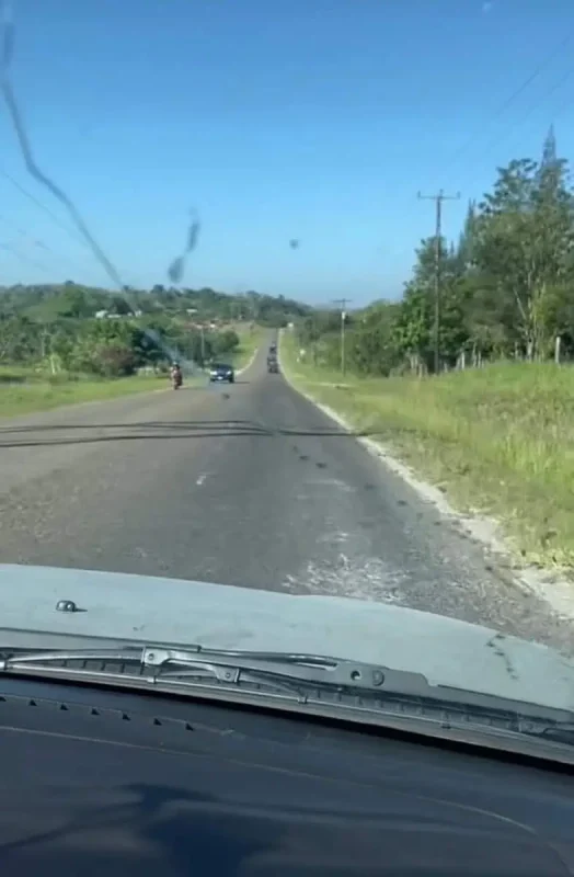 Travel San Ignacio To Flores & Crossing The Border 28 A long, straight road stretches into the distance through a lush green landscape, seen from inside a vehicle. Cars and motorcycles are visible on the road ahead, and the dashboard features windshield wipers and the edge of the windshield frame. The vehicle's shadow falls on the pavement as the road gently curves