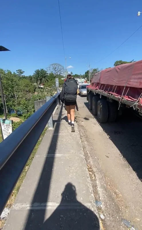 Travel San Ignacio To Flores & Crossing The Border 25 A female backpacker with a large black backpack and wearing a black tank top, shorts, and white socks walks on a narrow sidewalk beside a parked red tarp-covered truck when traveling from san ignacio to flores. The sidewalk is raised, adjacent to a guardrail overlooking a river, and leads through a small town with colorful buildings and clear skies.