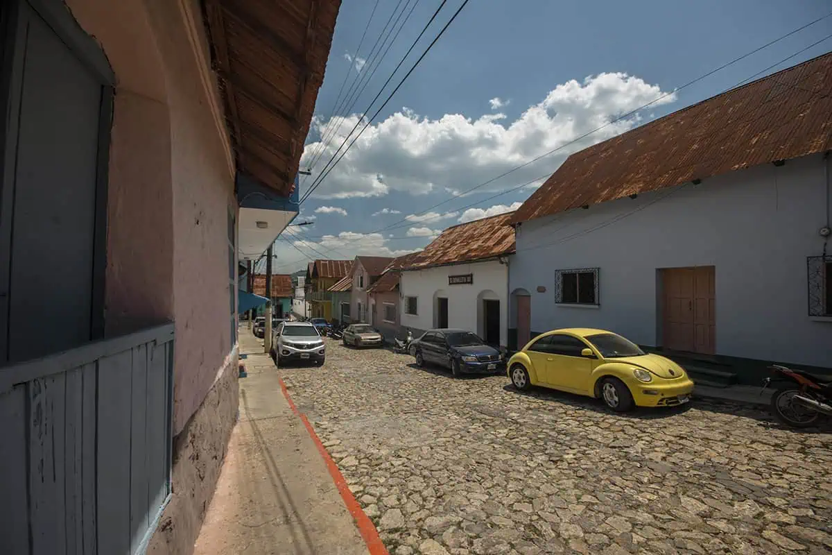 Travel San Ignacio To Flores & Crossing The Border 1 A cobblestone street lined with colorful buildings and parked cars after arriving from San Ignacio and Flores. The bright yellow VW Beetle stands out under the clear sky.