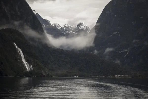 A breathtaking view of Milford Sound features the majestic Bowen Falls cascading down a lush green mountainside into the calm, reflective waters below. Snow-capped peaks rise in the background, shrouded in mist and low-hanging clouds. The contrast between the dark mountain slopes and the bright white snow creates a dramatic and ethereal scene.